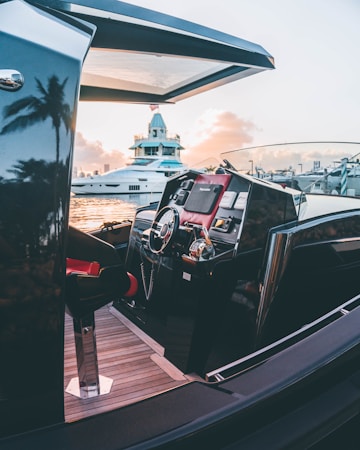 A luxurious yacht with a sleek, modern design is docked near a marina during sunset. The steering wheel and control panel of the yacht are visible, featuring polished metal and wood accents. The background shows another yacht and a building with a slightly vintage architecture. The scene is enhanced by the reflection of palm trees and the warm glow of the sunset.