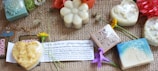 A collection of artisanal soaps arranged on a woven surface. The soaps are in various shapes such as hearts, flowers, and squares, with intricate designs embossed on them. Accompanying the soaps are small flowers and a card that likely explains the products. Tags with text like 'CHOOSE' and 'POP Starlit' are also present.