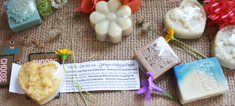 A collection of artisanal soaps arranged on a woven surface. The soaps are in various shapes such as hearts, flowers, and squares, with intricate designs embossed on them. Accompanying the soaps are small flowers and a card that likely explains the products. Tags with text like 'CHOOSE' and 'POP Starlit' are also present.
