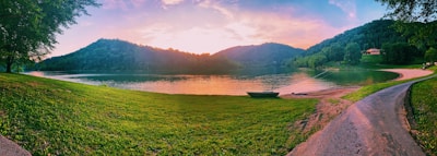A tranquil lakeside scene at sunset with a serene body of water surrounded by lush green hills. The sky displays a blend of warm colors. A small boat is docked near the shore, and a winding path leads towards a distant house nestled in the trees.