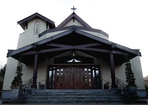 A church building with a large wooden door and steep staircase. The structure features a cross on the roof peak, arched windows, and is flanked by potted plants. The exterior walls are a light beige color, contrasting with the dark wooden trimmings of the roof and support beams.