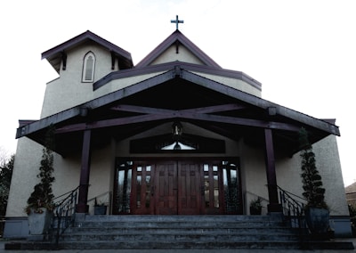 A church building with a large wooden door and steep staircase. The structure features a cross on the roof peak, arched windows, and is flanked by potted plants. The exterior walls are a light beige color, contrasting with the dark wooden trimmings of the roof and support beams.