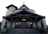 A church building with a large wooden door and steep staircase. The structure features a cross on the roof peak, arched windows, and is flanked by potted plants. The exterior walls are a light beige color, contrasting with the dark wooden trimmings of the roof and support beams.