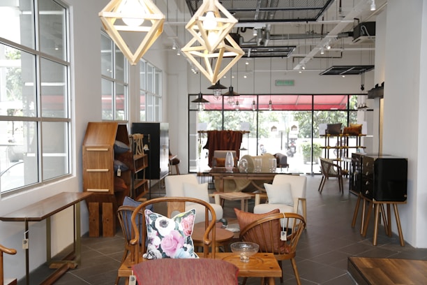An elegantly designed room features modern wooden furniture, with various types of seating and shelving. The space is well-lit due to large windows that allow natural light to stream in. Hanging geometric pendant lights add to the stylish atmosphere. The decor includes colorful throw pillows and minimal decorative pieces.