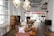 An elegantly designed room features modern wooden furniture, with various types of seating and shelving. The space is well-lit due to large windows that allow natural light to stream in. Hanging geometric pendant lights add to the stylish atmosphere. The decor includes colorful throw pillows and minimal decorative pieces.