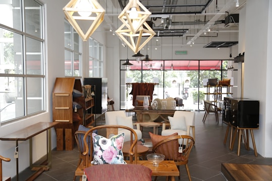 An elegantly designed room features modern wooden furniture, with various types of seating and shelving. The space is well-lit due to large windows that allow natural light to stream in. Hanging geometric pendant lights add to the stylish atmosphere. The decor includes colorful throw pillows and minimal decorative pieces.