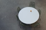 A round white table is surrounded by five green chairs, each with a unique, intricate design resembling branches or tree patterns. On the table, there is a single orange object, likely an orange or similar fruit. The surface on which the table and chairs sit is speckled and appears to be concrete or a similar material.
