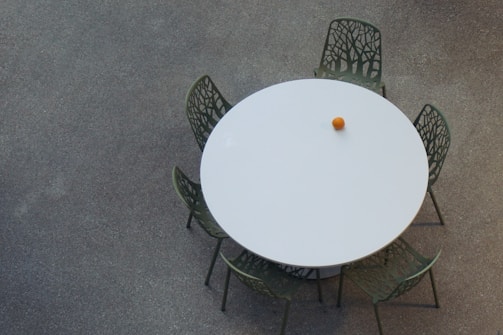 A round white table is surrounded by five green chairs, each with a unique, intricate design resembling branches or tree patterns. On the table, there is a single orange object, likely an orange or similar fruit. The surface on which the table and chairs sit is speckled and appears to be concrete or a similar material.