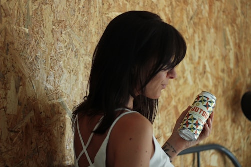 A woman with long dark hair is holding a can labeled 'Jutsu' with a colorful geometric design. She is wearing a white strappy top and standing against a textured wooden wall.