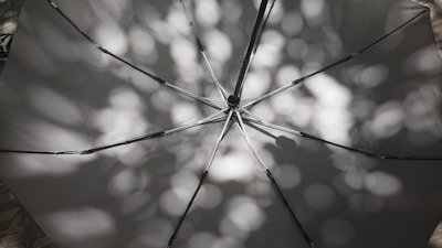 An open umbrella viewed from underneath, showing its skeleton and fabric. Dappled sunlight creates circular patterns of light and shadow on the fabric, adding a textured effect. The structure of the umbrella is clearly visible with metal rods radiating from the center.