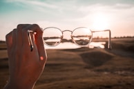 A hand holds a pair of round eyeglasses up against a backdrop of a sunset over a blurred landscape. The sunlight creates a halo effect around the glasses, and the reflection on the lenses captures part of the horizon and water.
