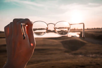 A hand holds a pair of round eyeglasses up against a backdrop of a sunset over a blurred landscape. The sunlight creates a halo effect around the glasses, and the reflection on the lenses captures part of the horizon and water.