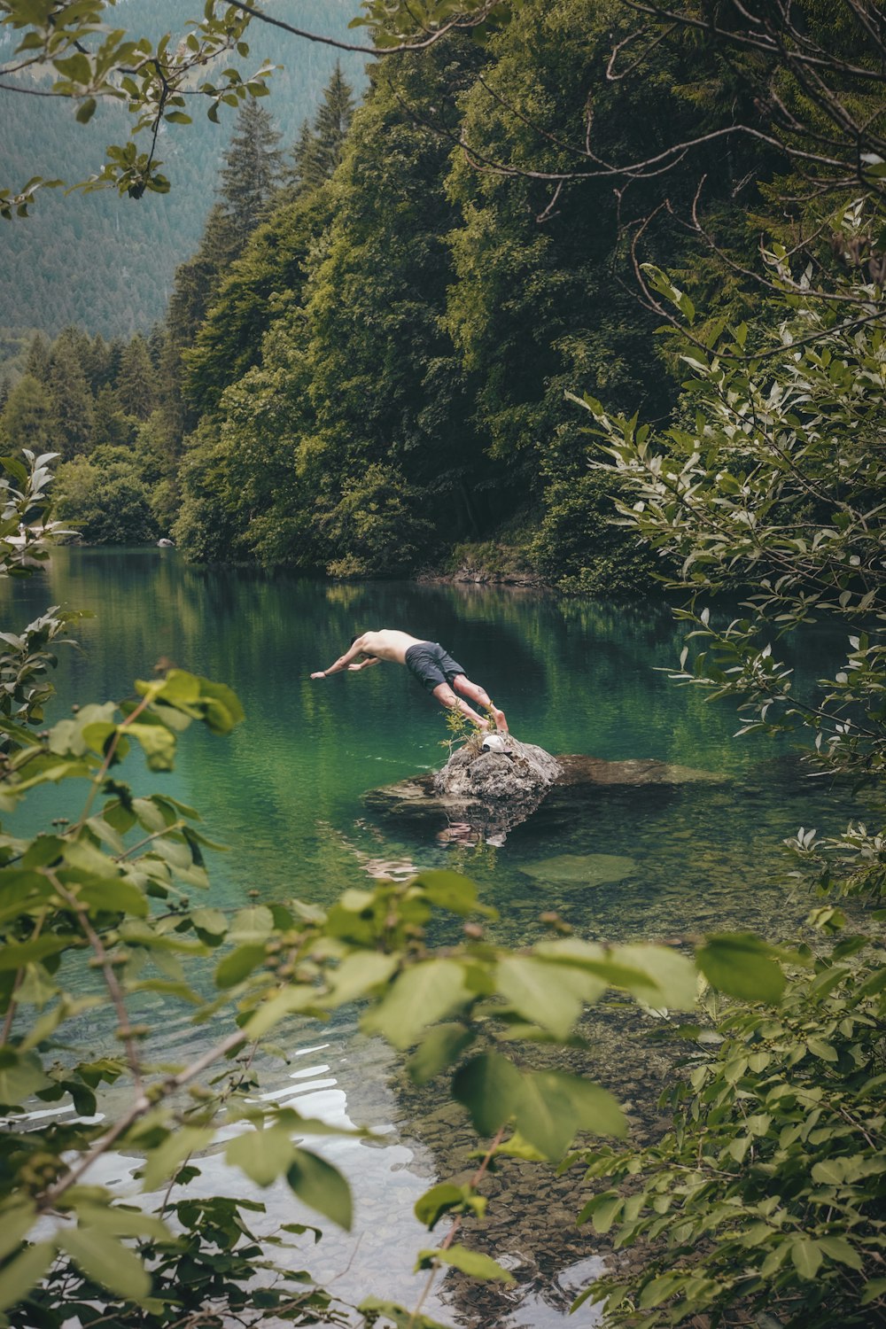 Hombre en topless con pantalones cortos negros a punto de zambullirse en el agua cerca de los árboles
