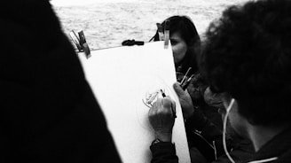 A monochrome image capturing a person sketching on a board while another person closely observes. The scene appears to be outdoors, possibly near water, as suggested by the textured background.