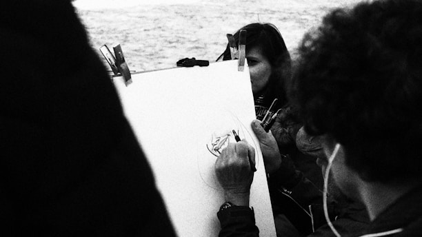 A monochrome image capturing a person sketching on a board while another person closely observes. The scene appears to be outdoors, possibly near water, as suggested by the textured background.