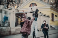 People are engaging in a playful activity near a building with a fence. One person is on stilts holding large cut-out numbers, while another is wearing a hat and reaching up. Confetti or snow-like particles are falling around them, suggesting a festive or celebratory mood.