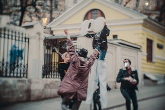 People are engaging in a playful activity near a building with a fence. One person is on stilts holding large cut-out numbers, while another is wearing a hat and reaching up. Confetti or snow-like particles are falling around them, suggesting a festive or celebratory mood.