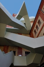 An abstract architectural structure featuring sharp, geometric lines and a combination of metal and brick materials. The design includes angular protrusions against a backdrop of a brick building and a clear blue sky.