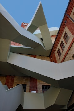 An abstract architectural structure featuring sharp, geometric lines and a combination of metal and brick materials. The design includes angular protrusions against a backdrop of a brick building and a clear blue sky.