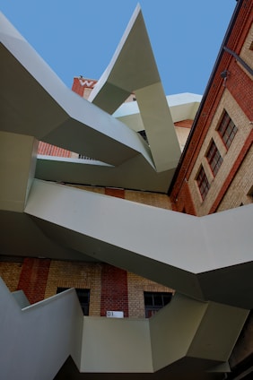 An abstract architectural structure featuring sharp, geometric lines and a combination of metal and brick materials. The design includes angular protrusions against a backdrop of a brick building and a clear blue sky.