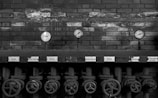 A series of industrial pressure gauges are mounted on a brick wall. Below them are several metal valves with handles, possibly part of an old machinery system. The image is in black and white, adding a vintage or historical feel.