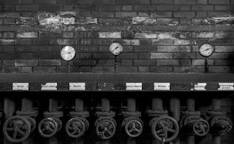A series of industrial pressure gauges are mounted on a brick wall. Below them are several metal valves with handles, possibly part of an old machinery system. The image is in black and white, adding a vintage or historical feel.