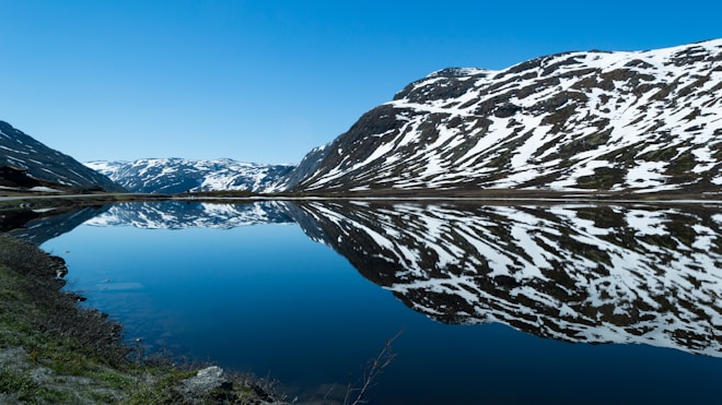 Snow-capped mountains are beautifully reflected in a tranquil, crystal-clear lake, creating a symmetrical illusion. The sky is clear and blue, enhancing the serene and pristine nature of the scene.