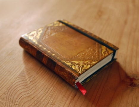 A vintage leather-bound book with intricate gold detailing on the cover is placed on a wooden surface. The book includes a red ribbon bookmark, peeking out from its pages.