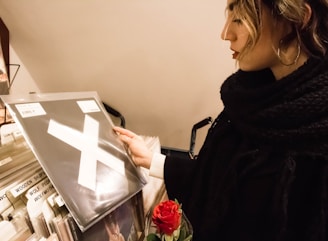 A person is browsing through a collection of vinyl records, holding a record with a large white 'X' on the cover. The individual is dressed in dark clothing and holds a red rose bouquet in their arm. The background consists of records in sleeves, displaying names like 'Wooden Ships' and 'Wolf People'.