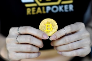 A person holds a golden coin with a Bitcoin symbol between their fingers. The background features the word 'REALPOKER' in bold letters on a dark shirt.