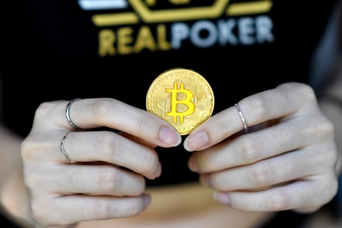 A person holds a golden coin with a Bitcoin symbol between their fingers. The background features the word 'REALPOKER' in bold letters on a dark shirt.