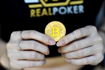 A person holds a golden coin with a Bitcoin symbol between their fingers. The background features the word 'REALPOKER' in bold letters on a dark shirt.