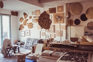 An artist's studio filled with organic and geometric art pieces hung on the walls. The room features a mix of textures, including intricately carved wooden art, framed drawings, and abstract decorations. A cluttered workspace with papers, tools, and materials is visible, along with a couch covered by a patterned throw. The light filters through a glass door on the left, creating a cozy and creative atmosphere.