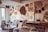 An artist's studio filled with organic and geometric art pieces hung on the walls. The room features a mix of textures, including intricately carved wooden art, framed drawings, and abstract decorations. A cluttered workspace with papers, tools, and materials is visible, along with a couch covered by a patterned throw. The light filters through a glass door on the left, creating a cozy and creative atmosphere.