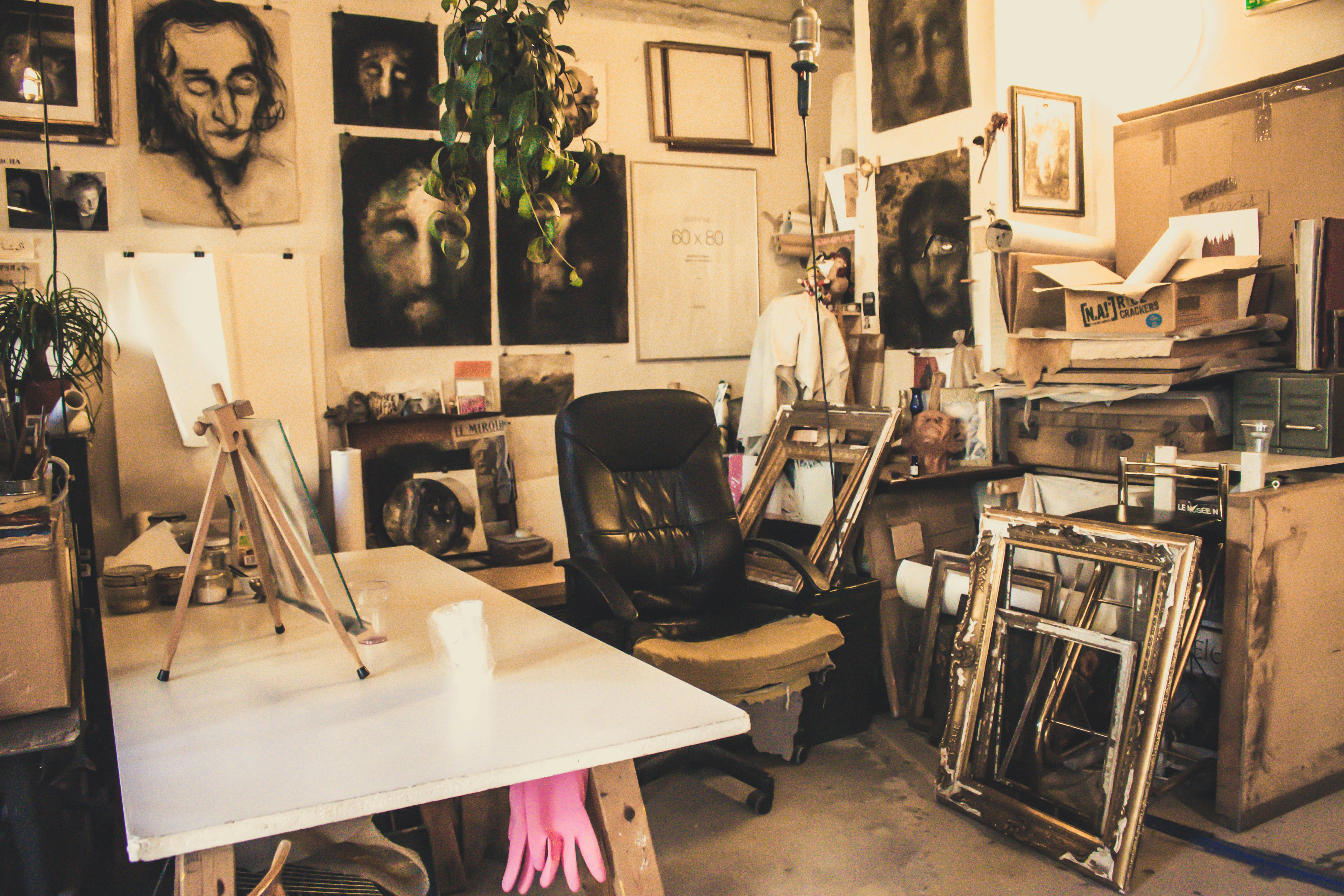 A cluttered artist's studio filled with canvases and paintings, some depicting human faces. An easel sits on a table with various art supplies scattered around. A black leather chair is placed in the center surrounded by wooden frames leaning against the walls. A plant hangs from the ceiling, adding a touch of greenery.
