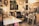 A cluttered artist's studio filled with canvases and paintings, some depicting human faces. An easel sits on a table with various art supplies scattered around. A black leather chair is placed in the center surrounded by wooden frames leaning against the walls. A plant hangs from the ceiling, adding a touch of greenery.