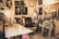 A cluttered artist's studio filled with canvases and paintings, some depicting human faces. An easel sits on a table with various art supplies scattered around. A black leather chair is placed in the center surrounded by wooden frames leaning against the walls. A plant hangs from the ceiling, adding a touch of greenery.