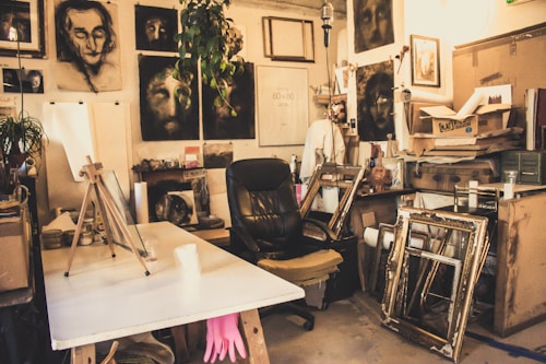 A cluttered artist's studio filled with canvases and paintings, some depicting human faces. An easel sits on a table with various art supplies scattered around. A black leather chair is placed in the center surrounded by wooden frames leaning against the walls. A plant hangs from the ceiling, adding a touch of greenery.