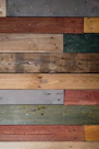 Various wooden planks are arranged horizontally, showcasing a range of colors and textures, including gray, brown, red, green, and beige. Some planks display visible screws and knots, giving a rustic and reclaimed appearance.