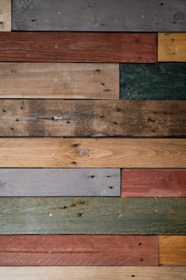 Various wooden planks are arranged horizontally, showcasing a range of colors and textures, including gray, brown, red, green, and beige. Some planks display visible screws and knots, giving a rustic and reclaimed appearance.