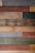 Various wooden planks are arranged horizontally, showcasing a range of colors and textures, including gray, brown, red, green, and beige. Some planks display visible screws and knots, giving a rustic and reclaimed appearance.