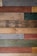Various wooden planks are arranged horizontally, showcasing a range of colors and textures, including gray, brown, red, green, and beige. Some planks display visible screws and knots, giving a rustic and reclaimed appearance.