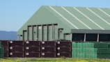 A large industrial warehouse with a green roof stands behind a stack of shipping containers. The containers are arranged in rows, with some in dark brown and others in green. The environment is sunny with a clear blue sky, and there are mountains visible in the background. The foreground features a patch of grass with yellow flowers.