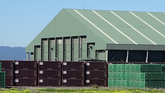 A large industrial warehouse with a green roof stands behind a stack of shipping containers. The containers are arranged in rows, with some in dark brown and others in green. The environment is sunny with a clear blue sky, and there are mountains visible in the background. The foreground features a patch of grass with yellow flowers.