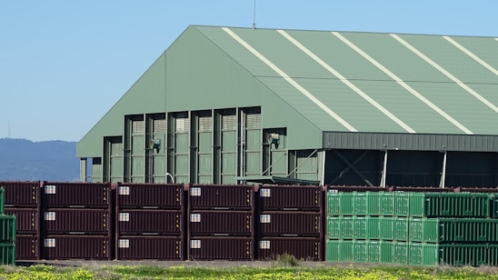 A large industrial warehouse with a green roof stands behind a stack of shipping containers. The containers are arranged in rows, with some in dark brown and others in green. The environment is sunny with a clear blue sky, and there are mountains visible in the background. The foreground features a patch of grass with yellow flowers.