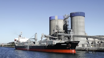 A large cargo ship is docked near industrial silos and a processing facility. The ship, named Kondili, has a black hull with a red lower section. Two sizable cylindrical silos with blue tops dominate the background, connected by various industrial structures and pipelines.