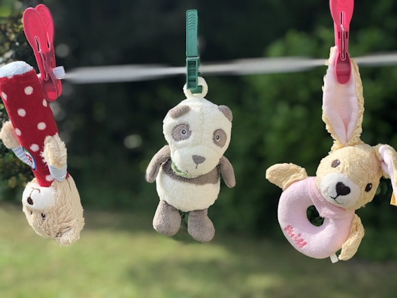 Three plush toys are clipped to a clothesline with plastic clothespins. The toy on the left appears to be a beige bear with a red polka dot outfit, hanging upside down by a pink clothespin. The center toy is a panda with a green clothespin. The toy on the right is a beige animal, possibly a bunny, with long ears and a pink ring around its neck, clipped by a pink clothespin. The background is a blurred green outdoor setting.