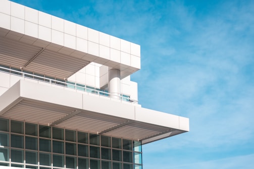 A modern building features clean, geometric lines with a focus on angular design. The structure is primarily constructed with white panels and has a series of horizontal and vertical beams. Large windows reflect the bright blue sky, adding a sense of openness.