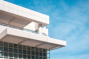 A modern building features clean, geometric lines with a focus on angular design. The structure is primarily constructed with white panels and has a series of horizontal and vertical beams. Large windows reflect the bright blue sky, adding a sense of openness.