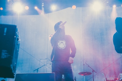 A performer wearing a hoodie is on stage, holding a microphone and singing. The lighting is bright and dramatic, with several beams and flares coming from stage lights above. Another person, also in a hoodie, stands to the right, partially visible. Music equipment, such as microphones and stands, is present in the background.